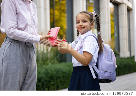 A mom hands her daughter a lunchbox at the schoolyard, preparing her for the school day. A mom hands her daughter a lunchbox at the schoolyard, preparing her for the school day. 129973587
