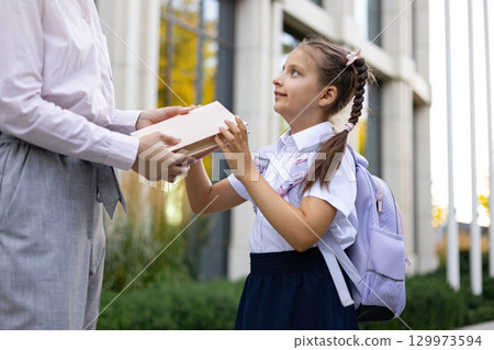 A mother hands her daughter a book outside a school building, capturing a moment of connection. A mother hands her daughter a book outside a school building, capturing a moment of connection. 129973594