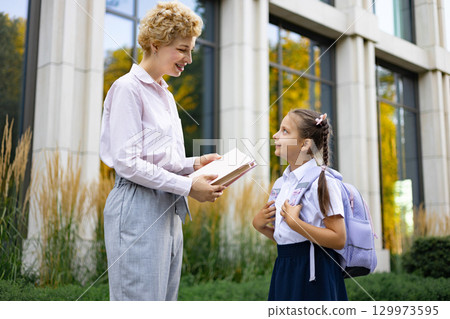 A mother smiles as she speaks to her daughter in a schoolyard, holding a book. A mother smiles as she speaks to her daughter in a schoolyard, holding a book. 129973595