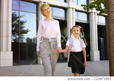 A mother and daughter walk hand in hand in front of a school building, a moment of connection and love. A mother and daughter walk hand in hand in front of a school building, a moment of connection and love. 129973665