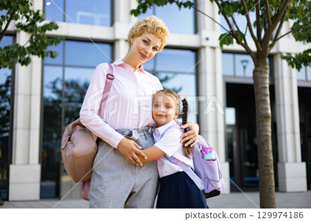A mother embraces her school-aged daughter, posing in front of a school building. 129974186