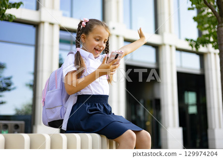 A young school girl with a backpack enthusiastically using her phone on a school campus. 129974204
