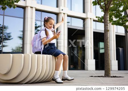 A young schoolgirl in a uniform sits on a bench, smiling while holding a phone, a backpack next to her, on a school yard. 129974205