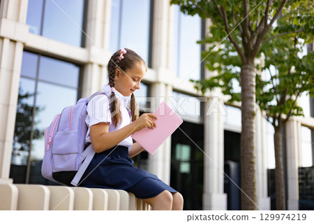 A young girl with braids and a backpack sits outside school, reading a pink book. A young girl with braids and a backpack sits outside school, reading a pink book. 129974219