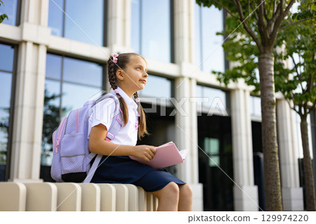 A young girl with a backpack sits outside a school building, holding a book and looking up. 129974220