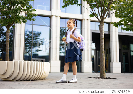 A young girl in school uniform stands outside a modern building, holding an apple and wearing a backpack. 129974249