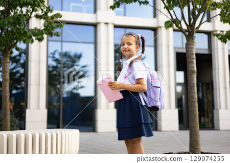 A schoolgirl with a backpack stands in front of the school building, holding a book. 129974255