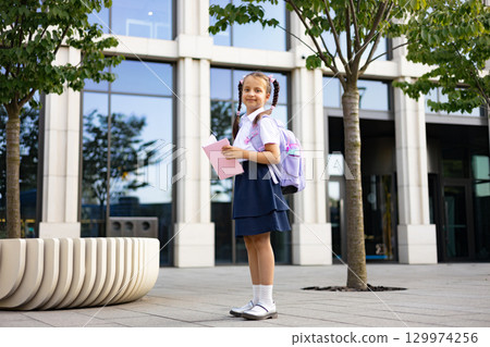 A schoolgirl with pigtails stands outside, holding a book and wearing a backpack, ready for her studies. 129974256