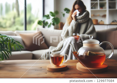 a cozy and minimalist scene with a glass teapot filled with amber-colored tea. In the background, there is blurred woman sits on sofa, wool scarf, cold blows her nose into a tissue, Livingroom, winter 129977084