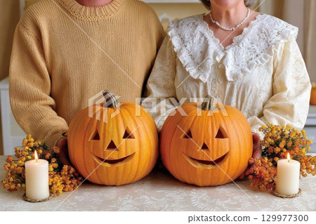 Elderly couple standing behind two carved pumpkins with smiling faces, surrounded by candles and autumn flowers, celebrating Halloween with cozy, festive decorations 129977300