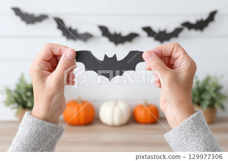 Elderly person holding a black paper bat decoration with pumpkins and bat garland in the background, preparing for Halloween in a bright and cozy indoor space 129977306