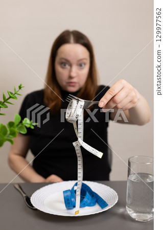 Person Shows Fork With Measuring Tape Instead of Food at Dining Table During Diet Reflection 129977562