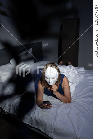 Person Wearing a Mask on Bed Using Smartphone in a Dimly Lit Room Focused on Privacy Protection Person Wearing a Mask on Bed Using Smartphone in a Dimly Lit Room Focused on Privacy Protection 129977587