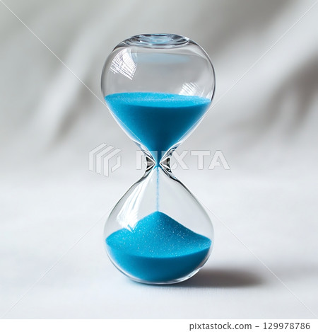 Hourglass with blue sand flowing down in front of white backdrop. 129978786