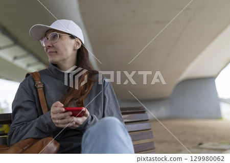 Woman using smartphone while sitting on bench under bridge 129980062