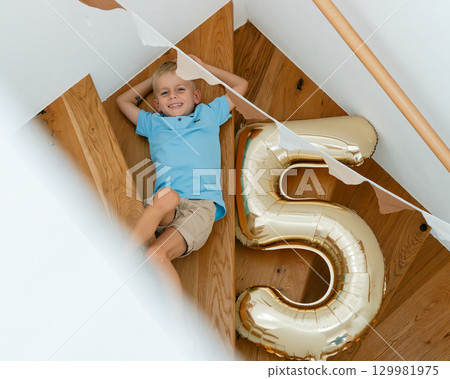 Young boy joyfully relaxing on wooden stairs with a large gold number five balloon, indicating his fifth birthday celebration. He wears a blue shirt and khaki shorts, smiling contentedly. 129981975