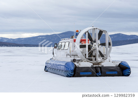 A hovercraft glides effortlessly over a snow-blanketed lake, surrounded by a serene winter landscape. The snow-covered surface reflects the cloudy sky, while distant mountains provide a picturesque A hovercraft glides effortlessly over a snow-blanketed lake, surrounded by a serene winter landscape. The snow-covered surface reflects the cloudy sky, while distant mountains provide a picturesque 129982359