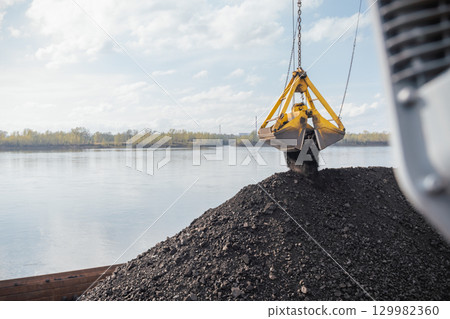Coal is being loaded at the port using a crane over the river during a sunny day, Coal is poured onto a barge against the background of a river from a bucket Coal is being loaded at the port using a crane over the river during a sunny day, Coal is poured onto a barge against the background of a river from a bucket 129982360