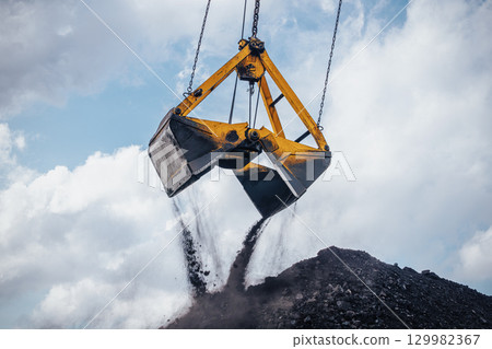 Heavy machinery is actively involved in unloading coal from a large bucket at an industrial location. The excavator crane, equipped with a dual clamshell bucket, hovers above a substantial pile of Heavy machinery is actively involved in unloading coal from a large bucket at an industrial location. The excavator crane, equipped with a dual clamshell bucket, hovers above a substantial pile of 129982367