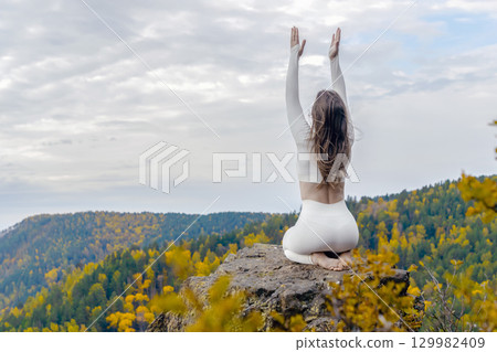 Woman sits on a rocky outcrop atop a mountain, engaged in yoga practice amidst vibrant autumn foliage. With her hands raised and back to the viewer, she embodies peace and focus in serene outdoors Woman sits on a rocky outcrop atop a mountain, engaged in yoga practice amidst vibrant autumn foliage. With her hands raised and back to the viewer, she embodies peace and focus in serene outdoors 129982409