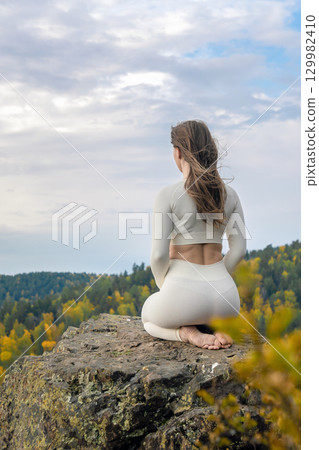 Meditation - A woman in yoga pose looks into the distance at the top of a mountain. Woman in white clothing practices yoga on a mountain during autumn, embracing nature and serenity 129982410