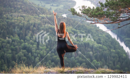 A woman in black practices yoga on a hilltop in nature outdoors against a background of green forest and river. Practicing yoga on a cliffside overlooking a lush green valley at sunset A woman in black practices yoga on a hilltop in nature outdoors against a background of green forest and river. Practicing yoga on a cliffside overlooking a lush green valley at sunset 129982411