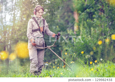 Woman using a lawn mower trimmer to maintain grass on a vibrant green lawn during daylight in a lush garden setting 129982438