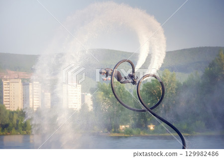 Flyboarding adventure showcasing acrobatics over calm water at sunset near city skyline. An athlete on a flytboard performs tricks over water. 129982486