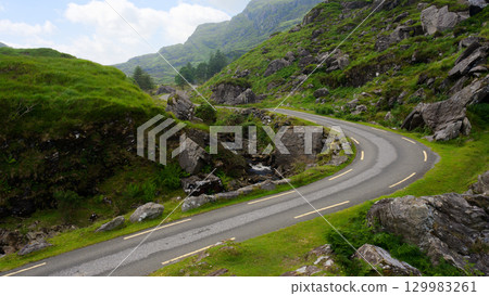 A winding road cuts through the picturesque Gap of Dunloe in Ireland. Green hillsides and rocky terrain surround the narrow asphalt road on a partly cloudy day. A winding road cuts through the picturesque Gap of Dunloe in Ireland. Green hillsides and rocky terrain surround the narrow asphalt road on a partly cloudy day. 129983261