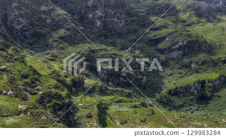 Grassy areas and rocky cliffs form the landscape of the Gap of Dunloe in Ireland. The green vegetation and rugged terrain create a striking contrast in County Kerry. Grassy areas and rocky cliffs form the landscape of the Gap of Dunloe in Ireland. The green vegetation and rugged terrain create a striking contrast in County Kerry. 129983284