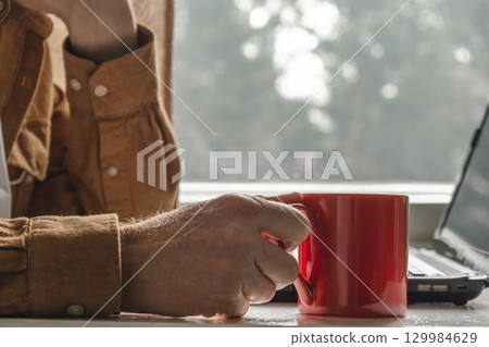 person holding red mug while working on laptop near window cozy soft natural light. closeup. person holding red mug while working on laptop near window cozy soft natural light. closeup. 129984629