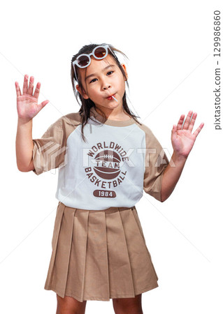 An Asian girl with sunglasses on her head eating a lollipop and raising both hands, posing playfully with a fun expression against a white background. An Asian girl with sunglasses on her head eating a lollipop and raising both hands, posing playfully with a fun expression against a white background. 129986860