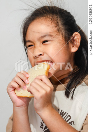 An Asian girl smiling while biting into a slice of bread, showing a playful and happy expression during mealtime. An Asian girl smiling while biting into a slice of bread, showing a playful and happy expression during mealtime. 129986861