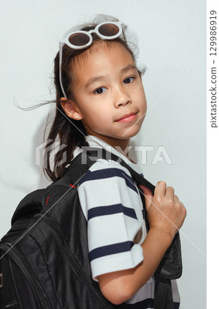 An Asian girl student with a backpack and sunglasses on her head, wearing a striped shirt and looking at the camera. 129986919