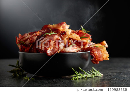 Fried bacon in a black dish with rosemary on a black background. Fried bacon in a black dish with rosemary on a black background. 129988886