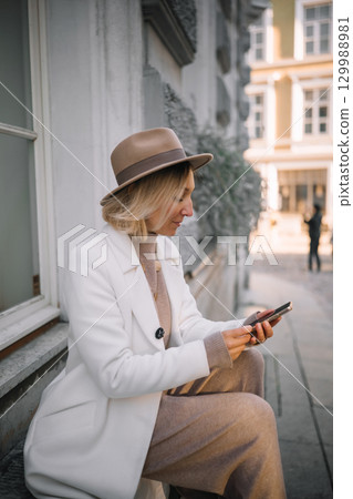 Stylish woman checks her phone while sitting on a sidewalk in a city 129988981