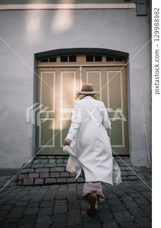Woman in white coat with hat walking toward vintage door on cobblestone street Woman in white coat with hat walking toward vintage door on cobblestone street 129988982
