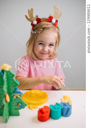 A young girl wearing festive antlers enjoys crafting holiday decorations at a table A young girl wearing festive antlers enjoys crafting holiday decorations at a table 129989011