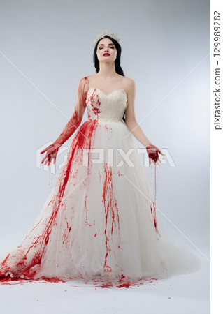 Stunning bride in elegant gown covered in blood stands with a knife 129989282