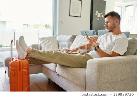 Man sitting on sofa with suitcase and using smartphone 129989720