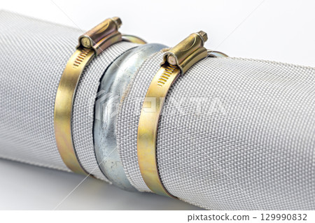 Metal pipe connector with brass strap, hose clamp, and flexible mesh hose, industrial equipment for plumbing or mechanical use, close up on white background 129990832