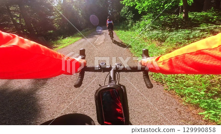 Point of view shot of a road cyclist riding behind a female cyclist on a sunny forest road, showing handlebars and bright red cycling jersey 129990858