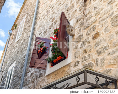 Rustic stone house with open shutters and floral baskets. Architecture, lifestyle, tradition and atmosphere of European village streets. 129992182