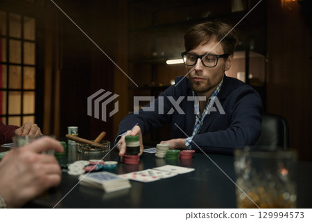 Stylish young man playing poker with his partners in a mysterious setting Stylish young man playing poker with his partners in a mysterious setting 129994573