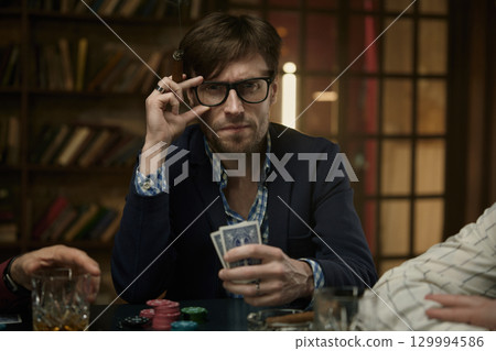 Man holding cards and a cigar at a poker table, surrounded by live poker game 129994586