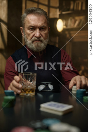 An experienced mature man sits at the poker table enjoying his whiskey and playing with chips 129994590