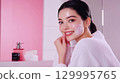 A smiling Asiatic woman enjoys a spa day, applying a hydrating face mask for healthy, glowing skin. A beautiful girl, lady with a radiant smile applies a facial mask This is AI-generated item 129995765