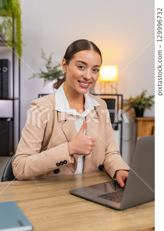 Young happy businesswoman shows thumbs up to camera confirms task complete on laptop smiles with joy Young happy businesswoman shows thumbs up to camera confirms task complete on laptop smiles with joy 129996732