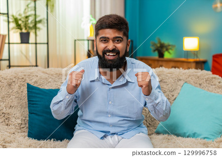 Indian man rising hands celebrates online game win happy news success great applause indoors victory Indian man rising hands celebrates online game win happy news success great applause indoors victory 129996758