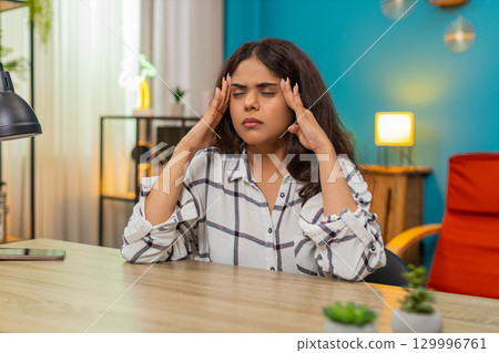 Indian woman holds head eyes closed grimacing pain strong severe headache health issue indoors home Indian woman holds head eyes closed grimacing pain strong severe headache health issue indoors home 129996761
